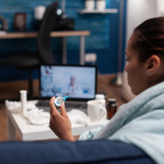 A young woman sick on the couch checking her temperature with a doctor on a laptop screen on the table