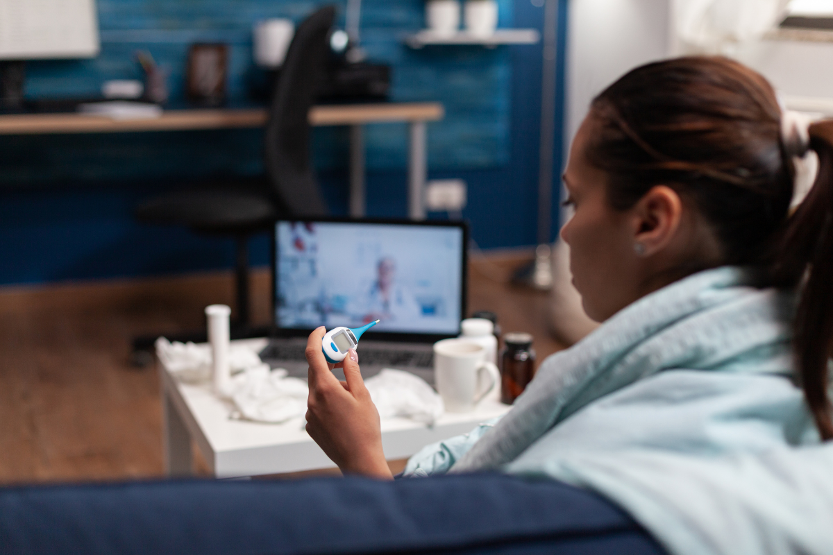 A young woman sick on the couch checking her temperature with a doctor on a laptop screen on the table