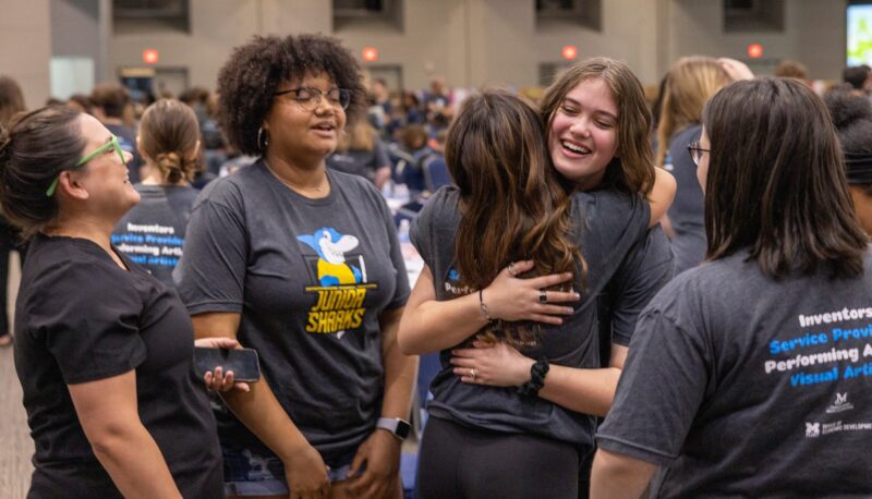 Two girls hugging while others look on.