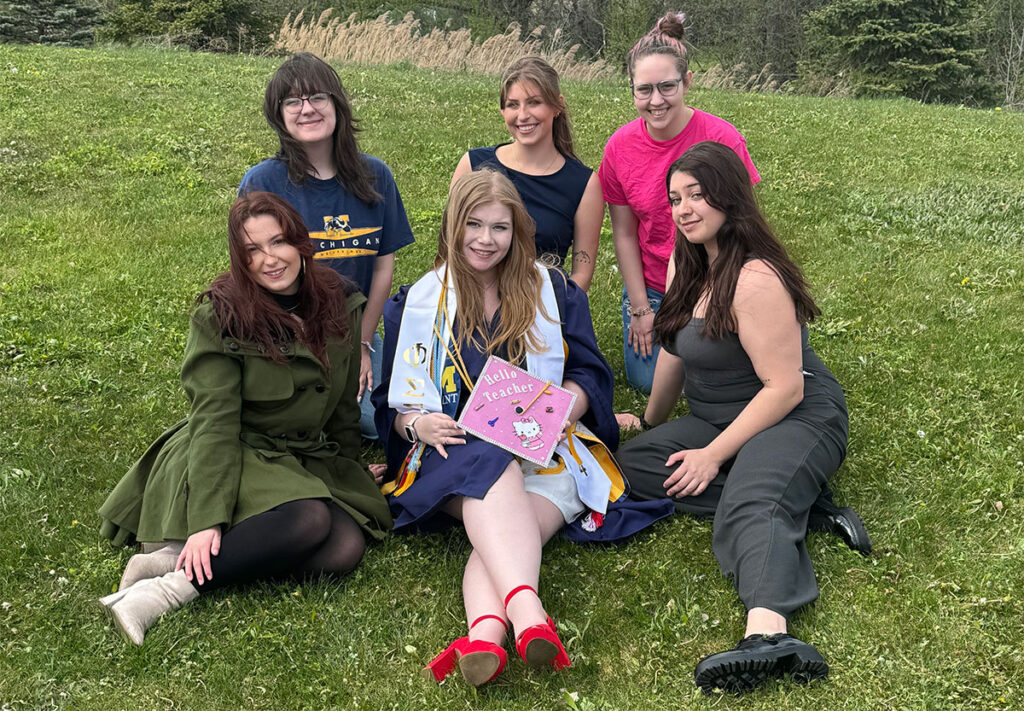 Six young, smiling women sit on a grassy hillside. The woman at the center is wearing a graduation gown and holding her cap, decorated in pink.