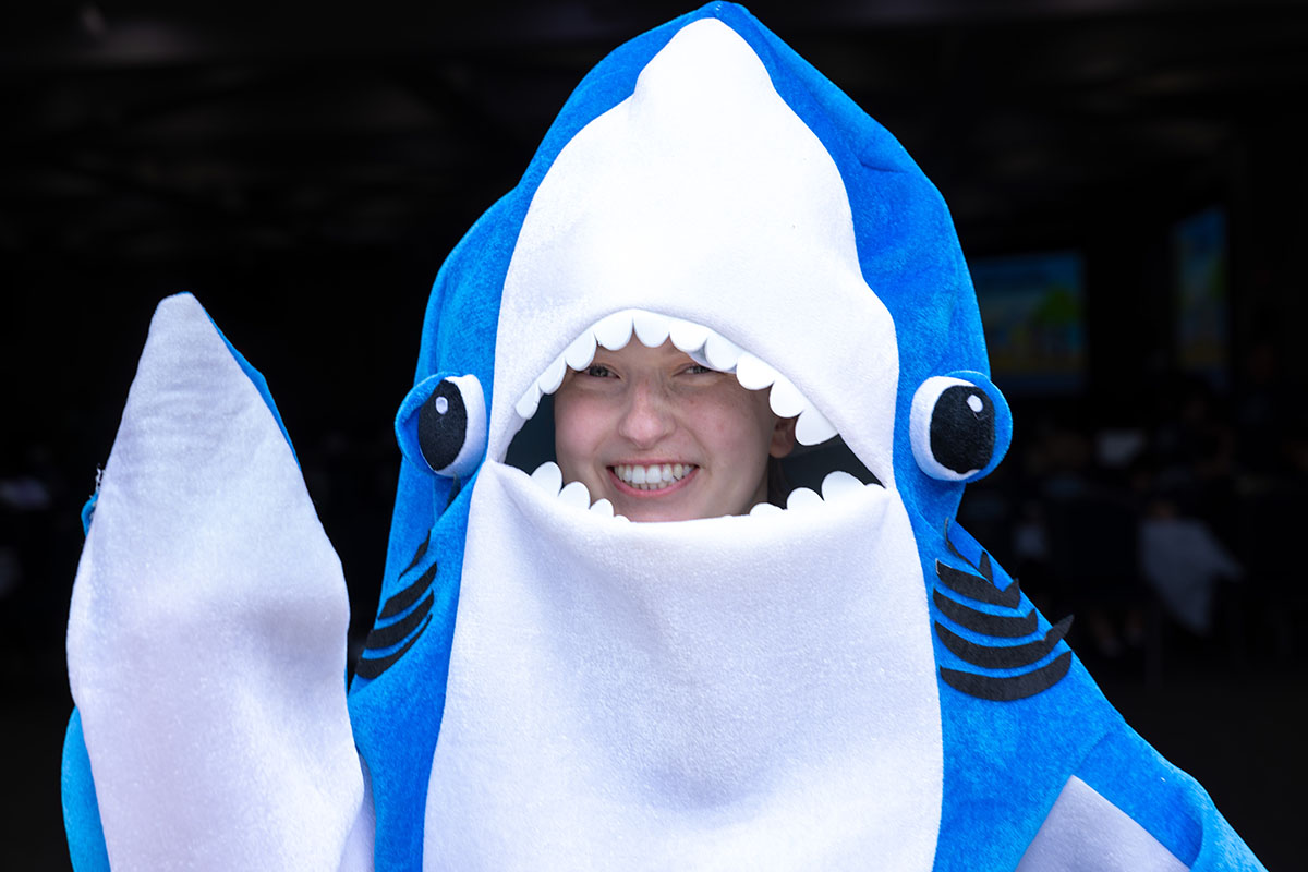 A person in a shark Mascot costume waving and smiling at the camera