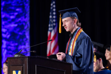 A young man in cap and gown speaking at a graduation ceremony