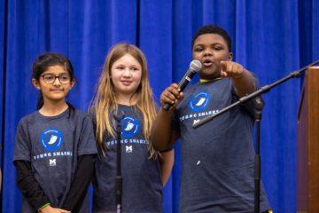 Three kids in Young Sharks t shirts standing on stage. A young boy is speaking into a microphone and pointing to the crowd, while two young girls look on.