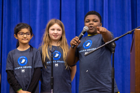 Three kids in Young Sharks t shirts standing on stage. A young boy is speaking into a microphone and pointing to the crowd, while two young girls look on.
