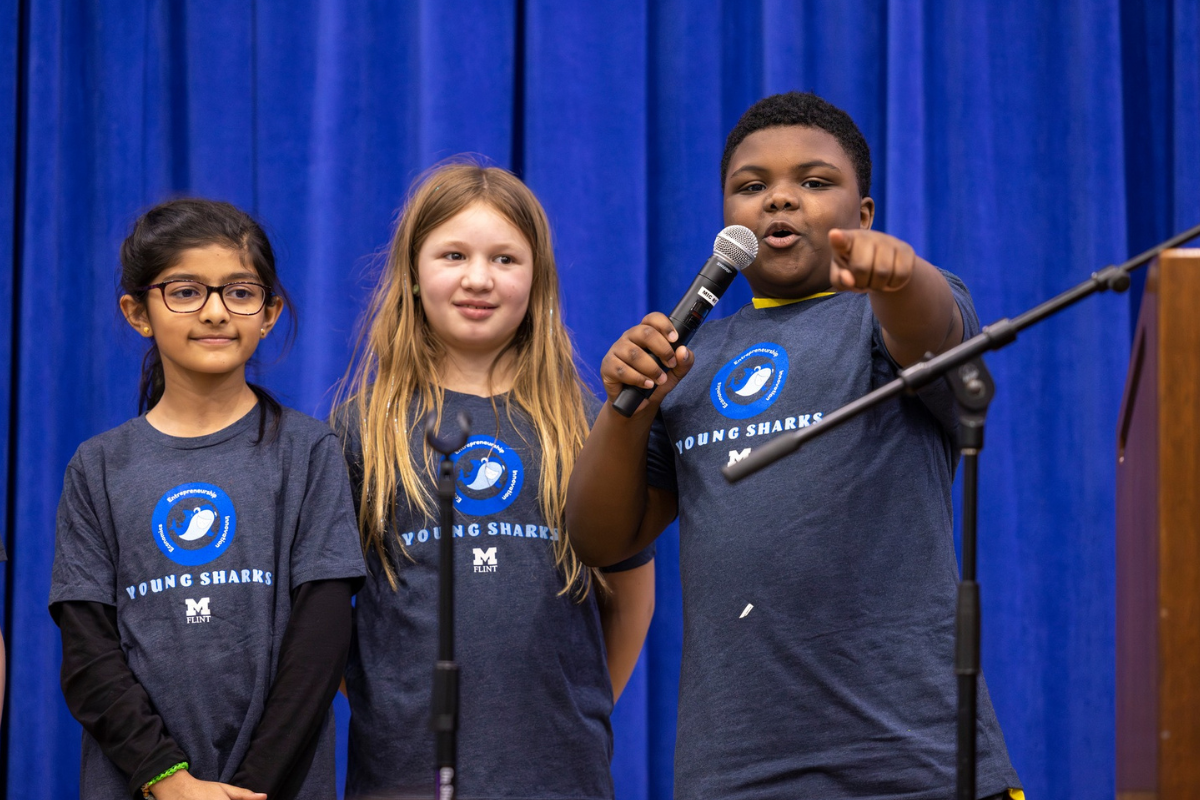 Three kids in Young Sharks t shirts standing on stage. A young boy is speaking into a microphone and pointing to the crowd, while two young girls look on.