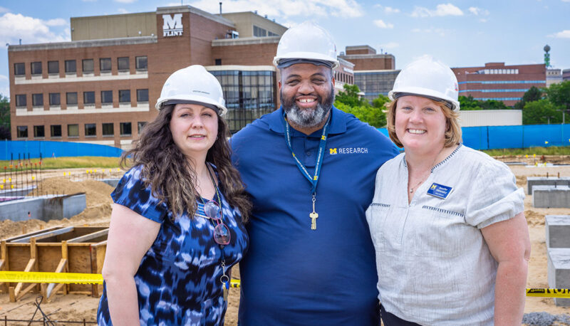 Three UM-Flint staffers posing with hard hats in front of the construction site.