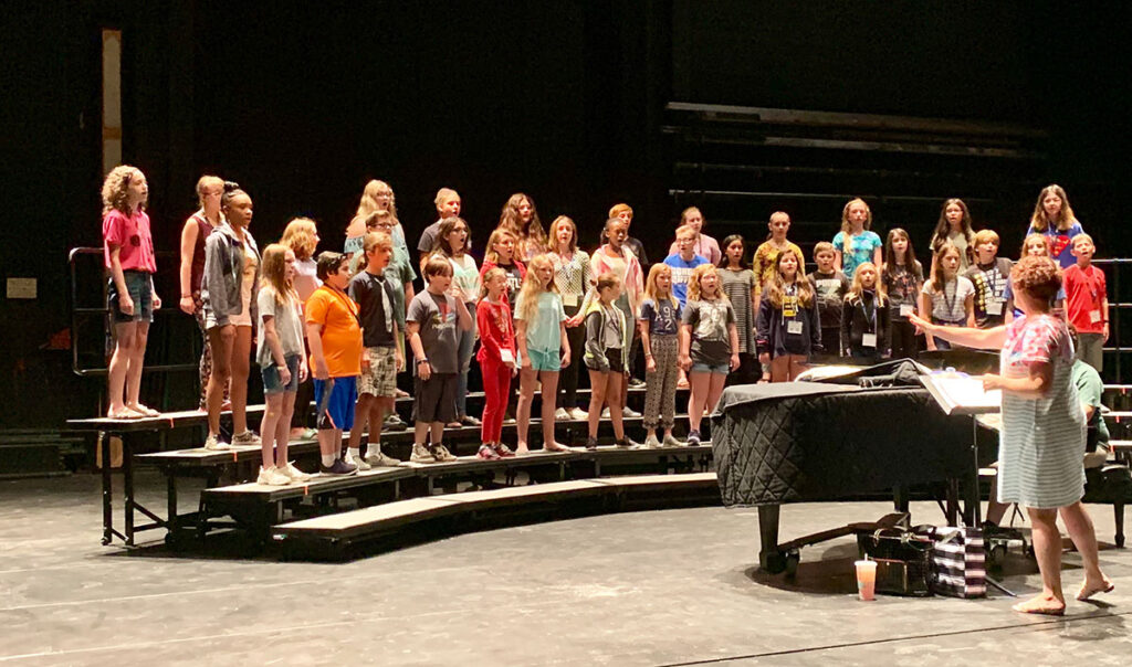 A group of children standing on risers practicing in a choir, with a teacher conducting them from behind a piano.