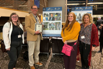 Thomas Henthorn posing with three students with the award in front of a poster detailing their cemetery work.