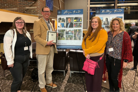 Thomas Henthorn posing with three students with the award in front of a poster detailing their cemetery work.
