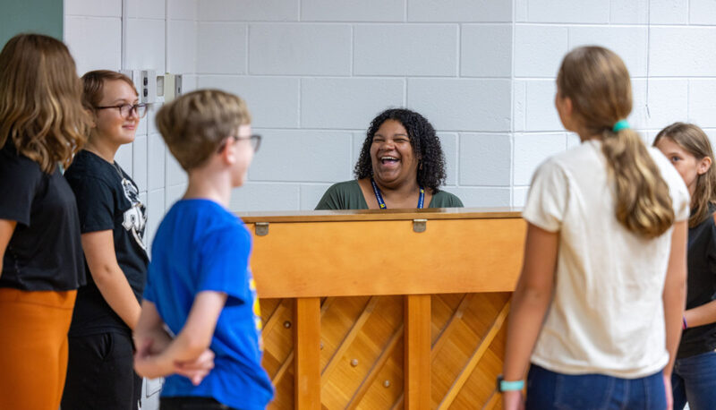 An instructor smiles while sitting at a piano while campoers look on.