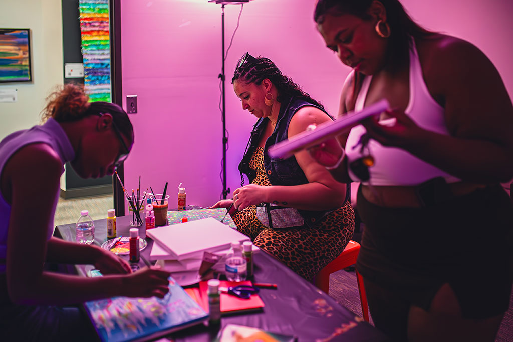 Three women sitting and standing around a table with canvases and art supplies, painting and making art in a purple-lit room with an art gallery in the background.