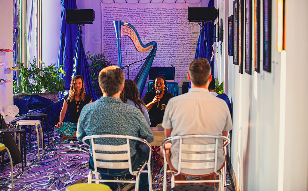 Several people sit in chairs in an art gallery with their backs facing us as two women sit on the floor, cross legged, one of whom is leading the group through a meditation practice.