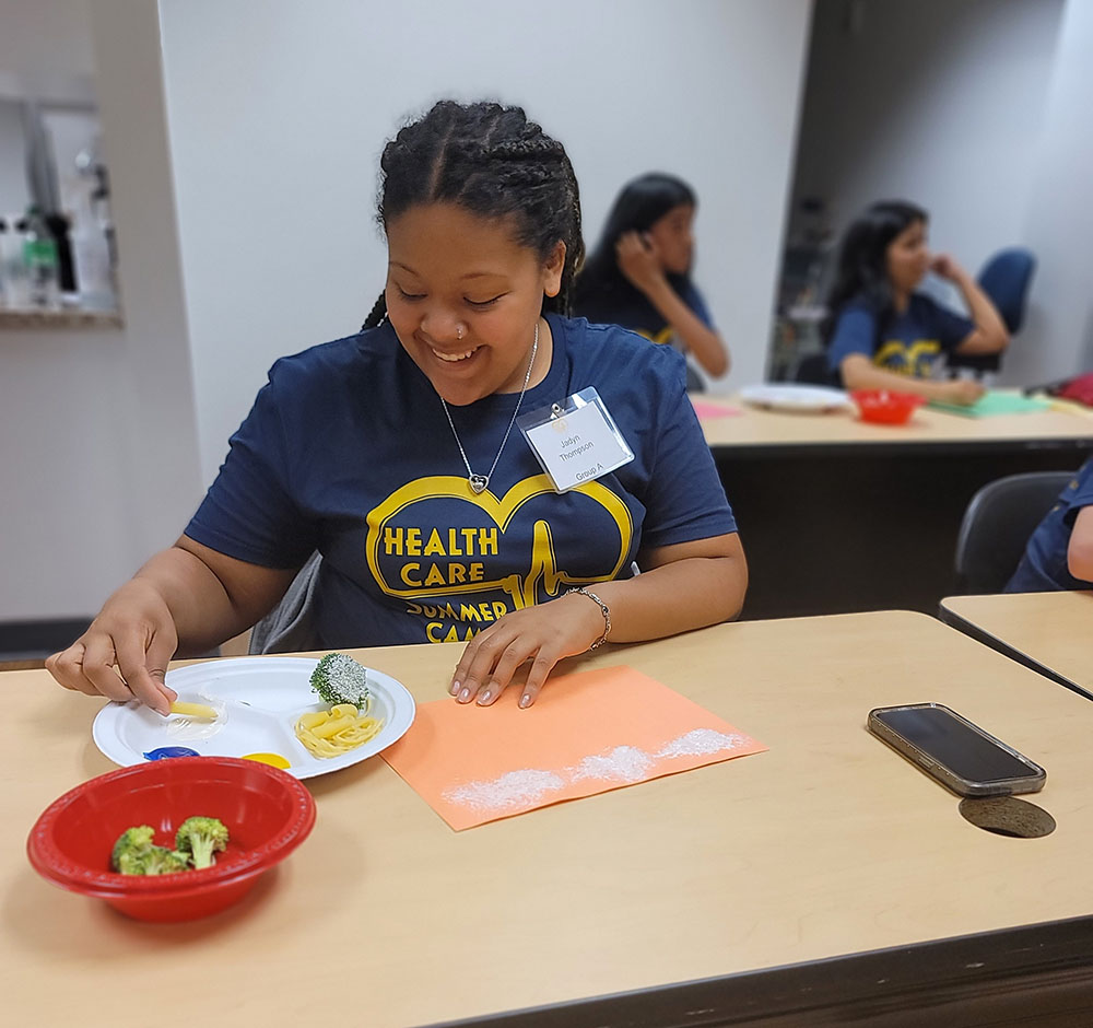 A camper is sitting at a desk taking prints of the textures different foods leave on paper after being dipped in glue. Among the foods are broccoli and dry pasta. 