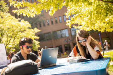 Two students sitting at a table outside. A male student in sunglasses is working on a laptop, while a female student reads a book.