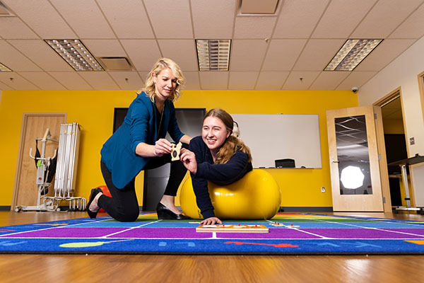 An OT student manipulating small objects while laying prone and balancing on a medicine ball, assisted by a faculty member. 