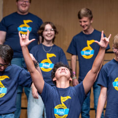 A singer thrusting his hands in the air with the "rock on" symbol with other campers on stage.