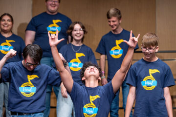 A singer thrusting his hands in the air with the "rock on" symbol with other campers on stage.