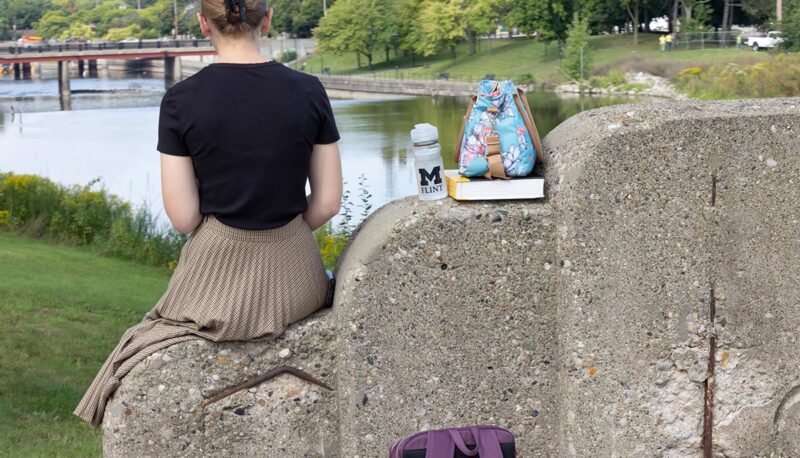 A student sitting on a concrete pillar looking at the Flint River with a UM-Flint water bottle.