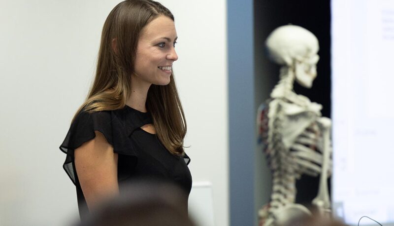 A lecturer at the front of a classroom. A model skeleton is also in view.