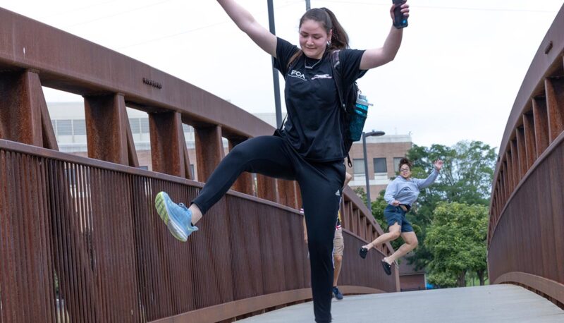 Students walking across the bridge on campus. They are jumping and clicking their heels for the camera.