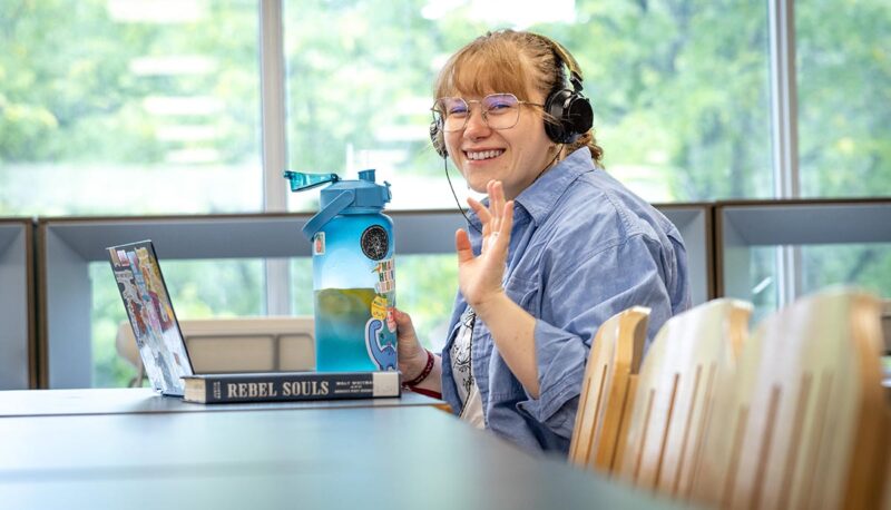 A student smiling and waving while working on a laptop in UCEN.