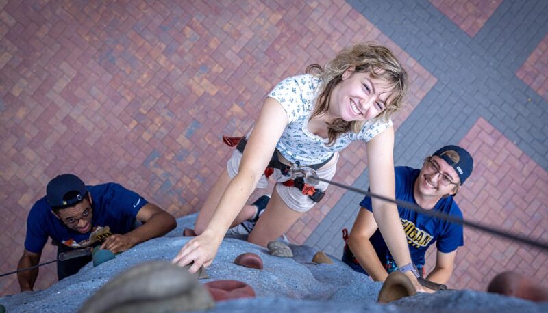 Three students on a climbing wall. The perspective is from the top of the wall down.