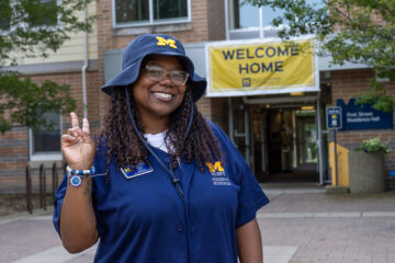 A UM-Flint staff member smiling and throwing a peace sign in front of Riverfront Residence Hall