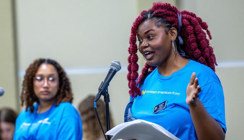 Jazlynne Cathey speaking at a microphone. She is a wearing a teal UM-Flint shirt and her hair is dyed red.