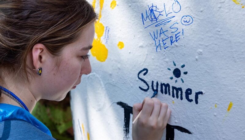 A student painting their name on the pillar. The name is "Summer" with a sun painted above it.