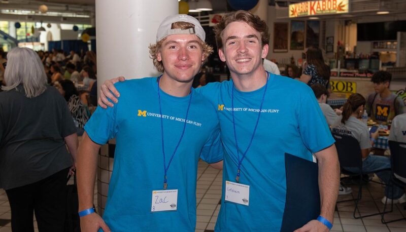 Two students in teal UM-Flint shirts smiling for the camera.