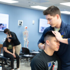 A physical therapy student manipulating the neck of another student. The same scene is playing out in the background of the photo.