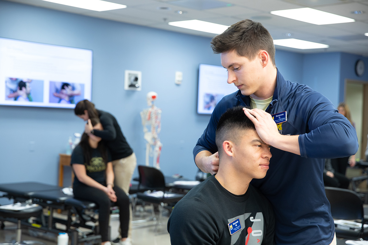 A physical therapy student manipulating the neck of another student. The same scene is playing out in the background of the photo.