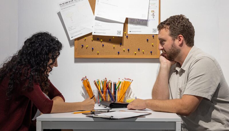 Gallery visitors seated at a desk writing their own UM-Flint memories.