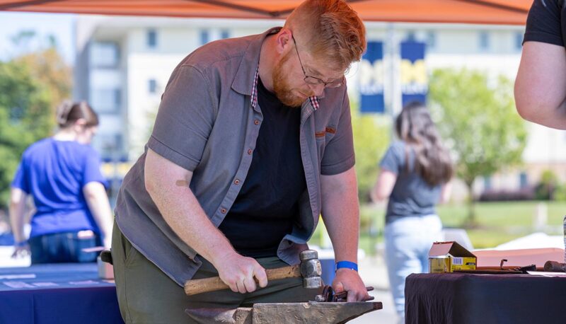 A student using a hammer and anvil under a tent.