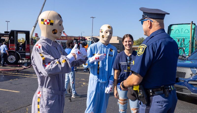 Two crash test dummy mascots talking to a state trooper