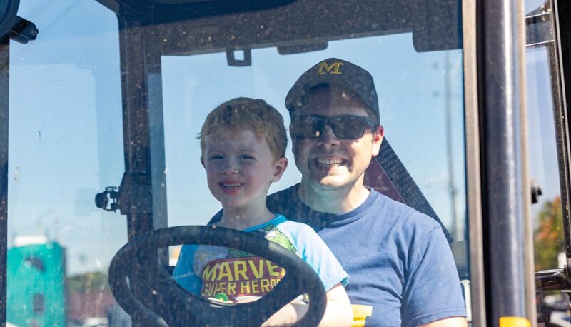 A young child sitting in the driver's seat of a big truck on his dad's lap.