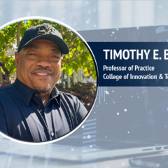 A photo of Timothy E. Bates, professor of practice at UM-Flint's College of Innovation & Technology, wearing a University of Michigan baseball cap against a backdrop displaying a laptop and a smartphone.