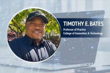 A photo of Timothy E. Bates, professor of practice at UM-Flint's College of Innovation & Technology, wearing a University of Michigan baseball cap against a backdrop displaying a laptop and a smartphone.