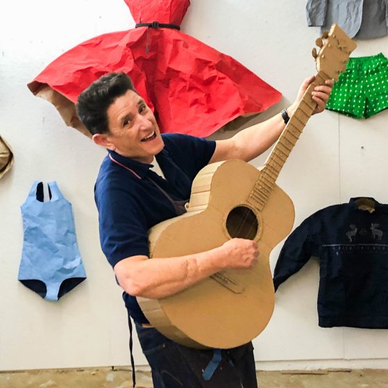 A photo of the artist Phranc holding an acoustic guitar and standing in front of her paper sculptures of clothing