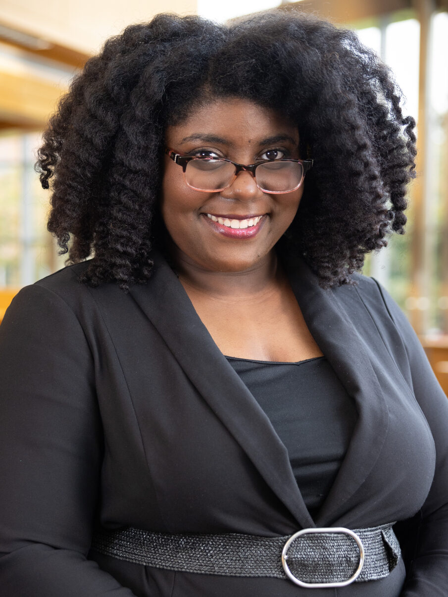 A portrait photograph of Alyssia Washington taken in a library. She is standing confidently, with her hands clasped in front of her, wearing a black dress.