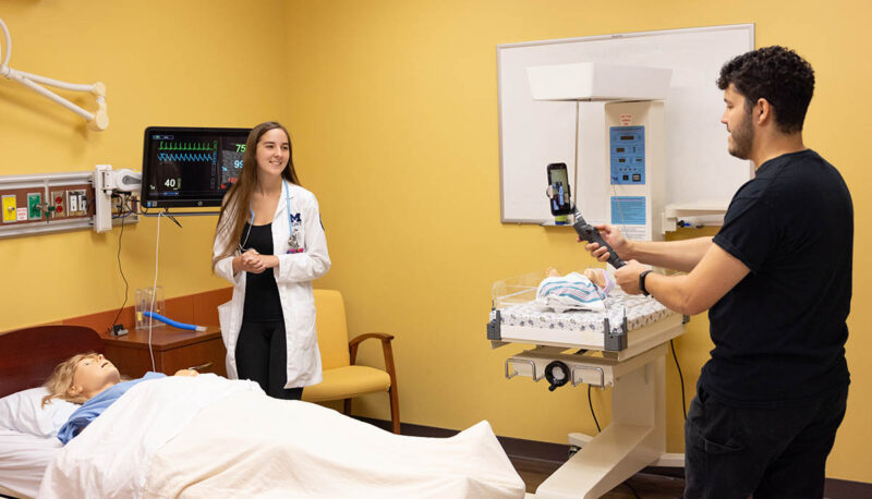 A health care student in a white coat being filmed in a simulated hospital room.