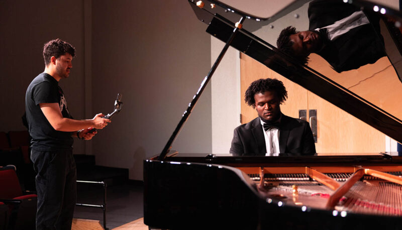 A student in a tuxedo playing the piano being filmed.