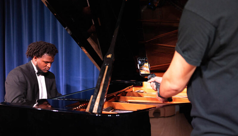 A student in a tuxedo playing the piano being filmed.