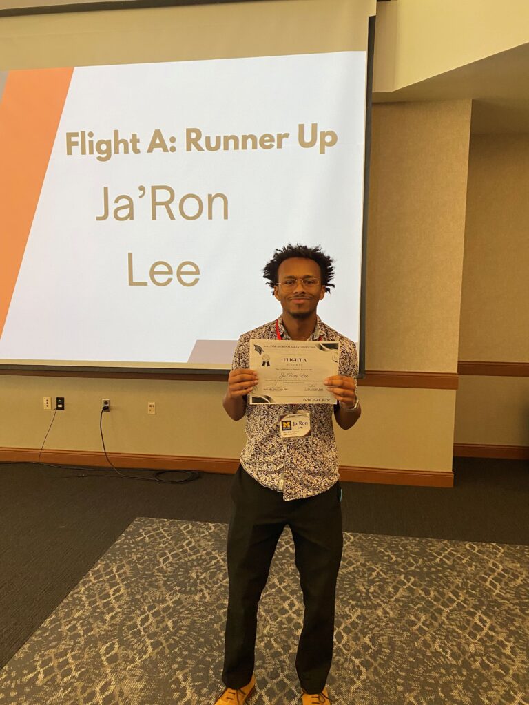 UM-Flint School of Management Marketing Student Ja'Ron Lee poses infront of a large screen holding his runner up award at the SVSU Regional Sales Competition on Oct. 11-12. 