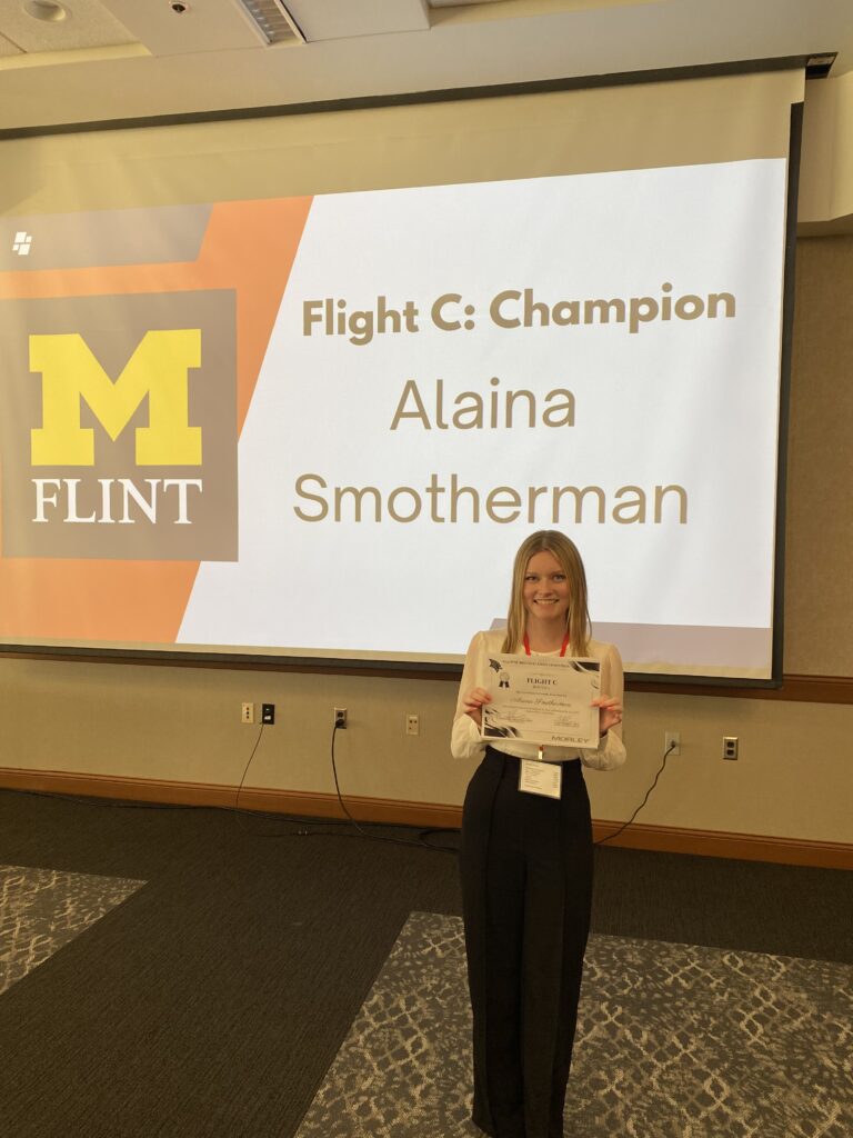 UM-Flint School of Management Marketing Student Alaina Smotherman poses infront of a large screen holding her champion award at the SVSU Regional Sales Competition on Oct. 11-12.