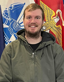 A portrait photograph of Christopher Mayes standing in front of various military flags.