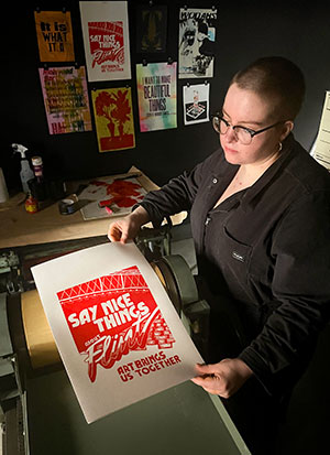 A photo of an artist holding a print that she has made that says 'Say Nice Things About Flint'