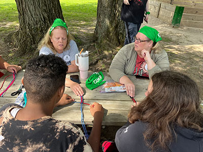 Two women wearing green handkerchiefs in their hair work with two campers to create woven multi-colored bracelets