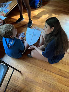 An intern sitting on the floor with a camper doing arts and crafts. 