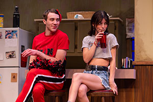 Two students sitting side-by-side forming. One is wearing a cheerleader outfit while the other is wearing cutoff jean shorts and a cutoff tank top. 